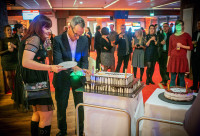 Two people cut into a large cake while people celebrate in the background.