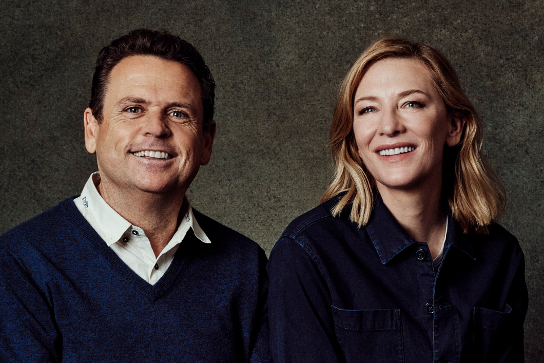Cate Blanchett and Danny Kennedy sit side-by-side against a grey background. They are looking at the camera and smiling. Both are wearing dark blue, Blanchett in a collared blue button down and Kennedy in a blue v-neck sweater over a cream button down shirt with small fish patterned on it. 