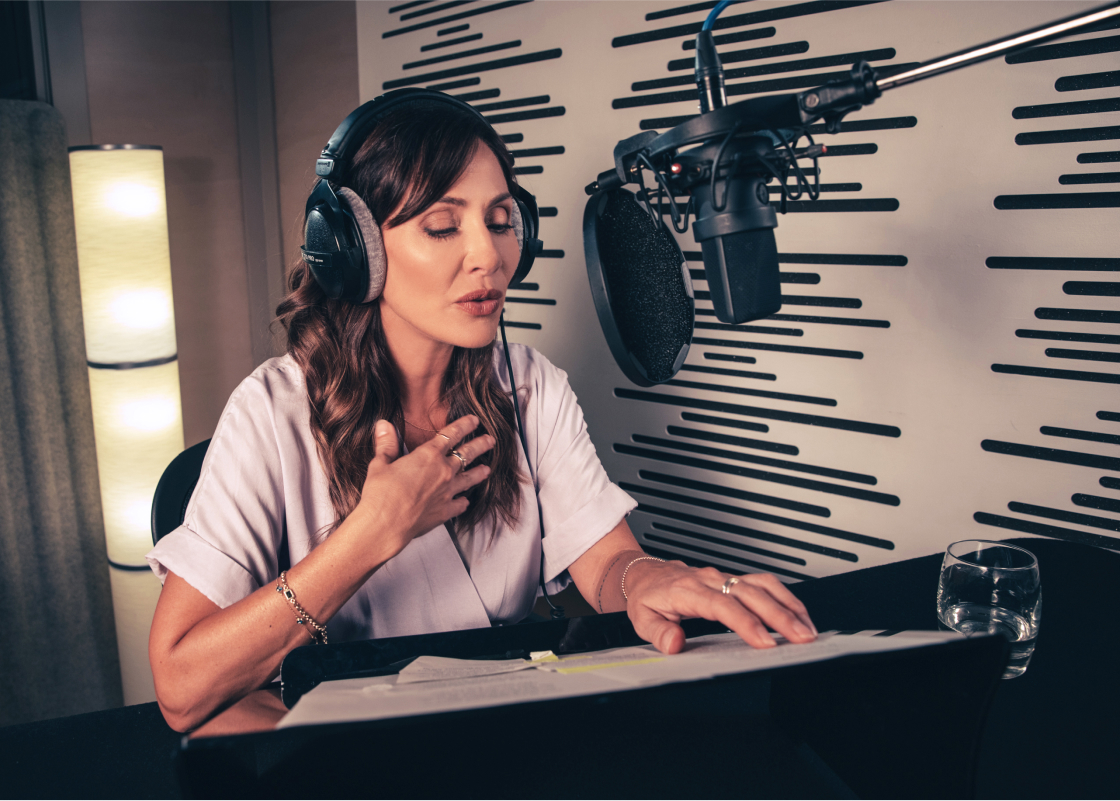 Natalie Imbruglia Sits in a recording studio in front of a microphone, a script open in front of her, and headphones on her head.