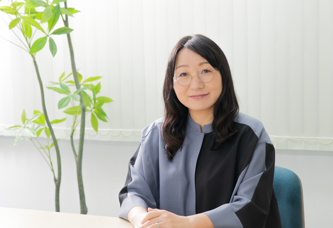 Saori Horikawa is seated at a desk, looking directly at the camera. She is wearing glasses and a gray and black shirt. Behind her is a large potted plant. 