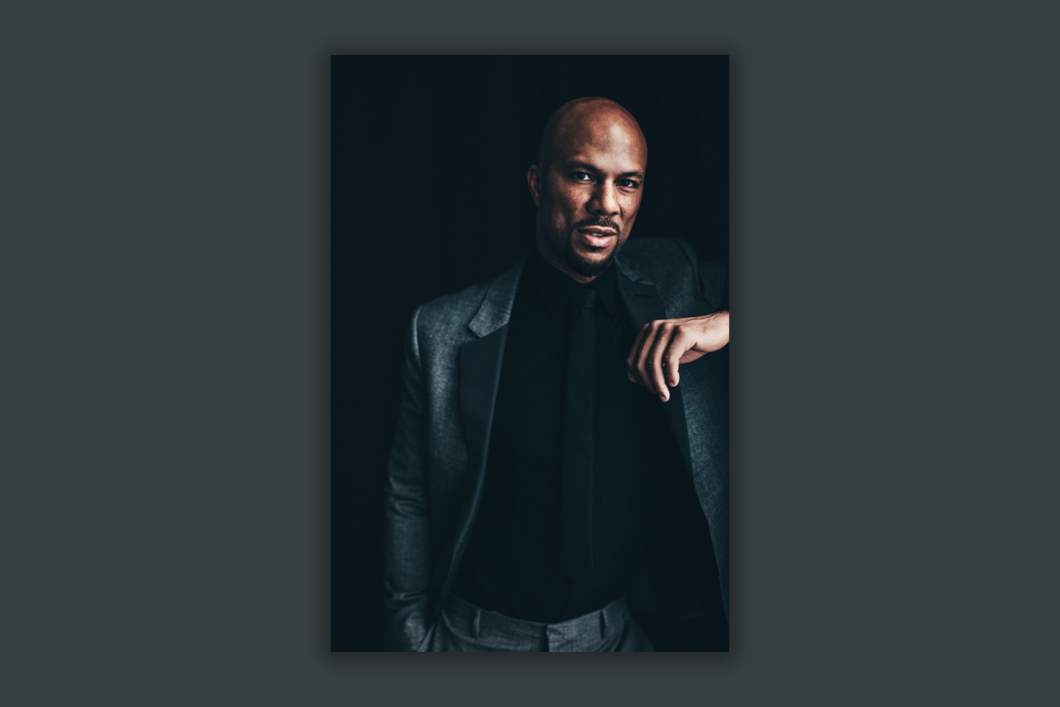 Oscar, Grammy, Emmy, and Golden Globe-winning artist, actor, activist and two-time New York Times best-selling author, Common, is wearing a black leather coat and black shirt. He is leaning to his left and is situated in front of a black background.