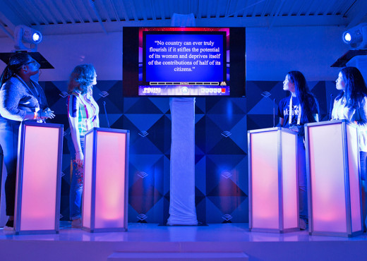 Two teams of women stand behind pink-lit podiums and face off against each other to answer a trivia question posted to a screen between them.