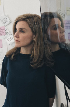 A woman with long blonde hair and with a black crew neck shirt on leans against a wall, her face reflected in the screen next to her, looking off in the distance with a wall of post-it notes behind her. 