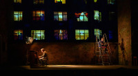 On a theatre stage, Jake Gyllenhaal sits playing a piano downstage right with a large light on him while Tom Sturridge sits on a rolling ladder upstage left looking out at the audience. An image is projected on the wall behind them to make it look like an apartment building with lots of lit windows framed by curtains. 