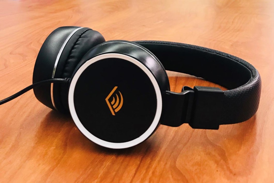 A pair of headphones rest on a wooden table top. The headphone are black and the over-the-head variety. On the outside of the cushioned earphones there is the orange chevron from the Audible logo.