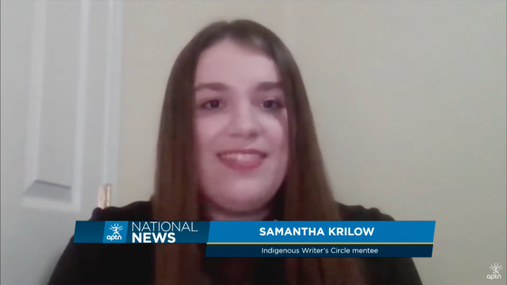 Indigenous Writers' Circle mentee Samantha Krilow is shown in news footage of an interview with her at home talking to the reporter via video. Her long brown hair is straight and she is wearing a dark shirt while sitting in a room with cream walls.
