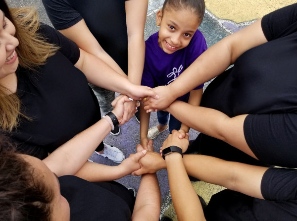 A group of Audible employees stand in a circle with a little girl during a volunteer event, their arms crossed over each other to form a ring of held hands at the circle's center. The photo is taken from the top down so the adult volunteers are seen as black t-shirts and hands and partial faces while the girl is clear at the top of frame and looking up and smiling. She is, frankly, adorable.