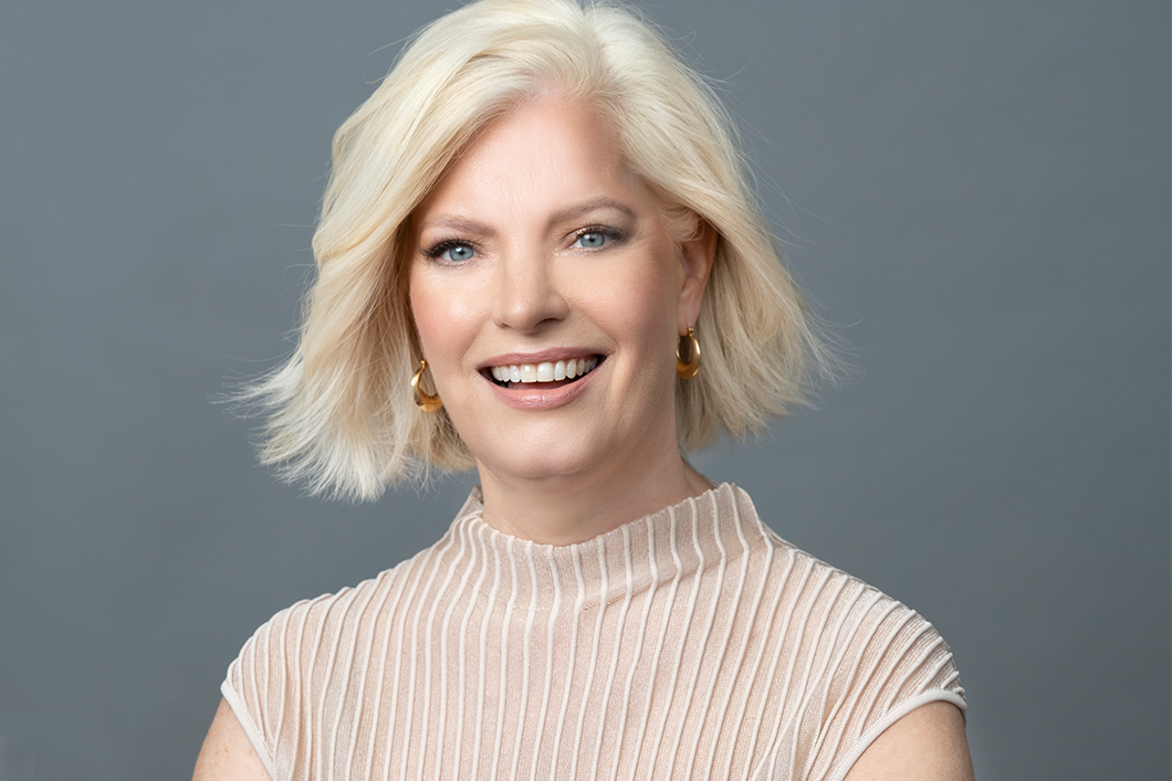 A headshot of Susan Jurevics, Audible's EVP, Head of International. Her head and shoulders are shown in close up, against a light gray background. She is looking directly to camera, smiling and wearing a light rose and white sweater. 