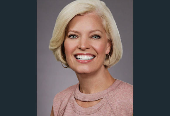 A headshot of Susan Jurevics, Audible's EVP, Head of International. Her head and shoulders are shown in close up, against a light gray background. She is looking directly to camera, smiling and wearing a light rose and white sweater. 