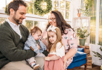 Audible employees Jessica and Joe Reese sit on a blue velvet couch in front of a picture window decorated with a holiday wreath holding their two small children in their laps. They are looking down at their children and smiling.