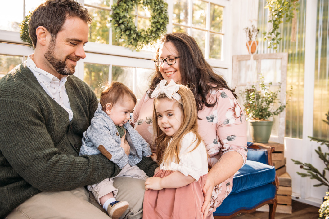 Audible employees Jessica and Joe Reese sit on a blue velvet couch in front of a picture window decorated with a holiday wreath holding their two small children in their laps. They are looking down at their children and smiling.