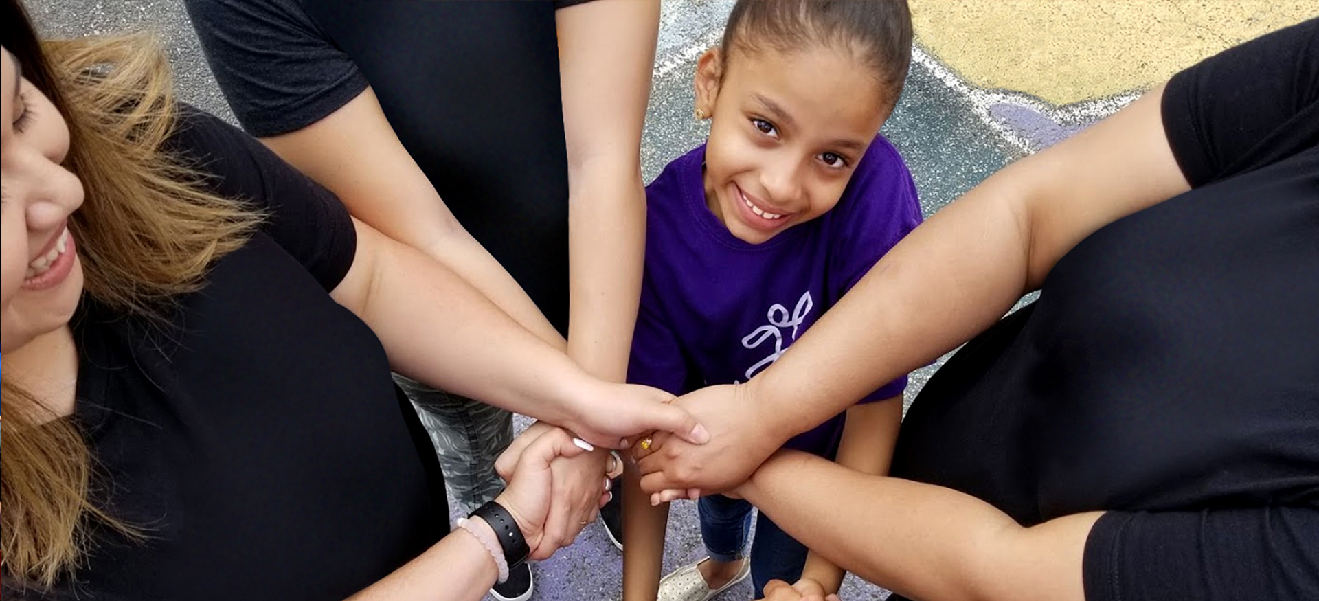 A group of Audible employees stand in a circle with a little girl during a volunteer event, their arms crossed over each other to form a ring of held hands at the circle&#x27;s center. The photo is taken from the top down so the adult volunteers are seen as black t-shirts and hands and partial faces while the girl is clear at the top of frame and looking up and smiling. She is, frankly, adorable.