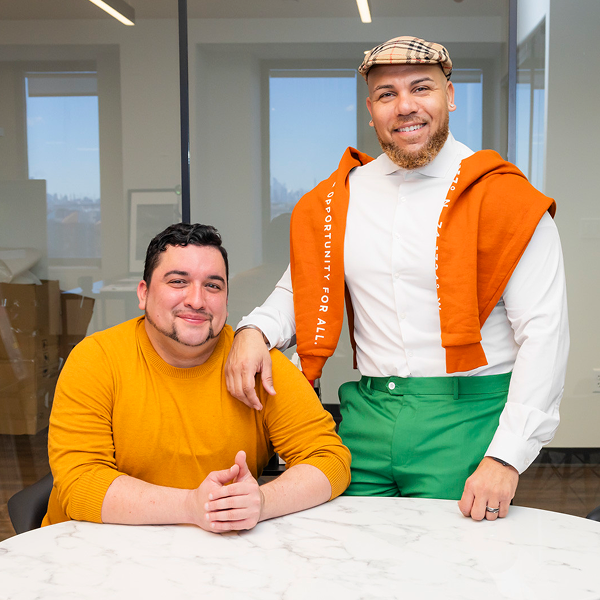 Two business partners pose for a portrait, one seated at a table, the other with his arm propped on the seated man's shoulder.