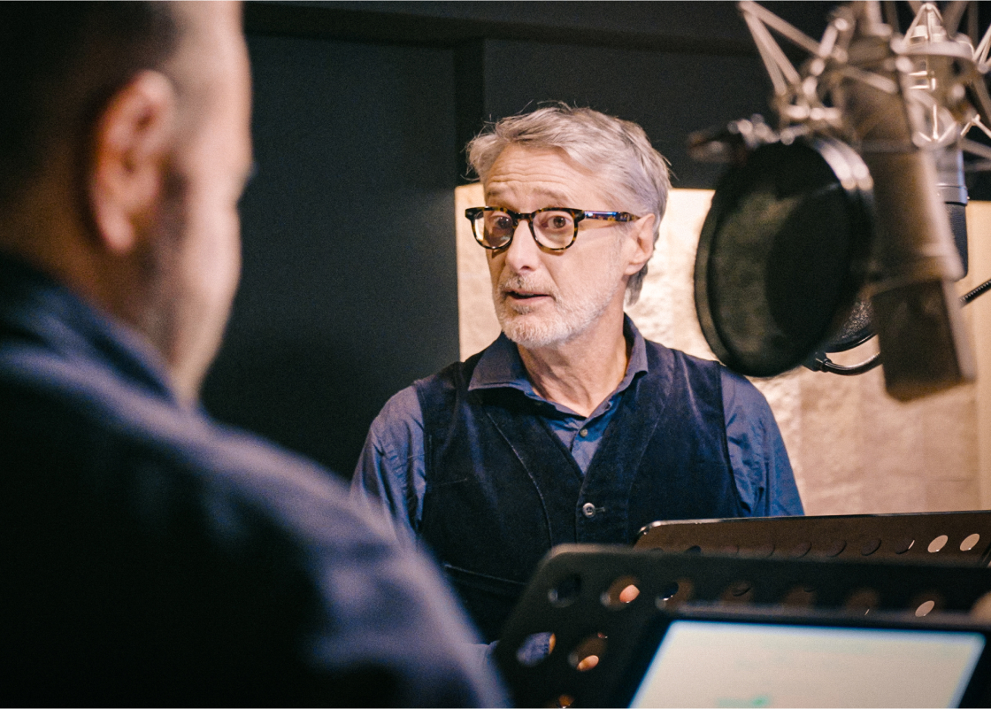 Antoine de Caunes stands at a microphone in a recording studio narrating an audiobook.