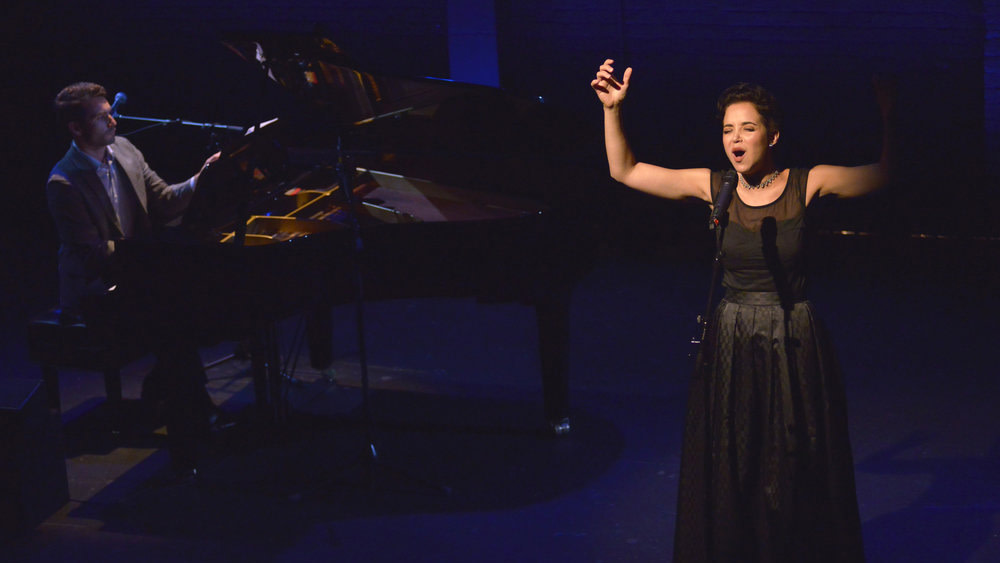 Alexandra Silber, wearing a black dress, sings at a microphone center stage. A man is playing a baby grand piano behind her.