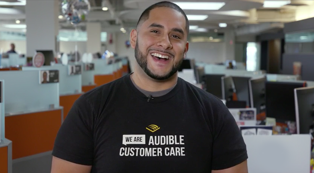 A smiling Raphael stands in Audible Customer Care center wearing a customer care shirt.