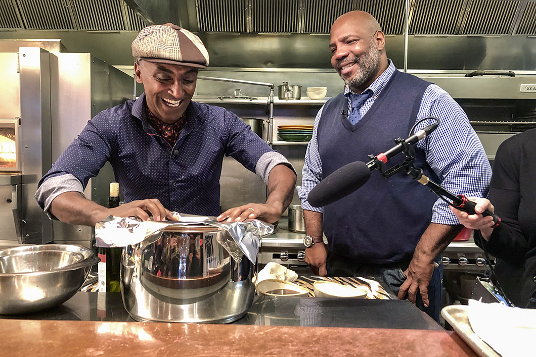 Chef Marcus Samuelsson at the stove of one of his restaurants with a guest standing by him smiling. 