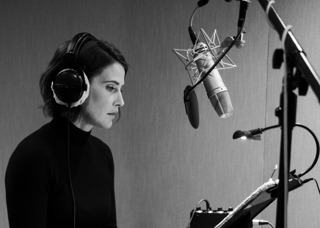 Colbie Smulders sits in a recording studio at a microphone performing an audiobook.