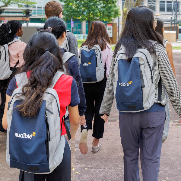 A few students are walking down the street with grey and blue backpacks on that show the Audible logo.
