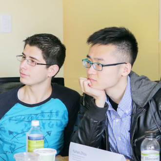Two interns sit side-by-side at a table paying intense concentration to a speaker off screen. They both have cropped hair and are wearing glasses.