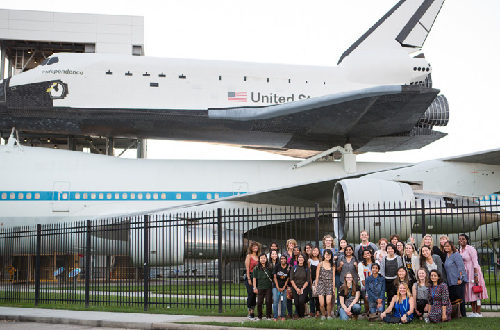 A large group of women are posed together in front of a fence. The space shuttle looms behind them.