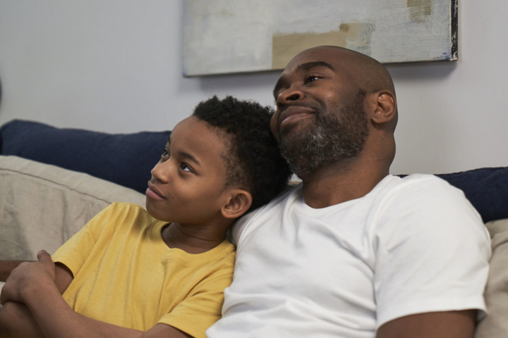 A man sits on a couch with his arm around his son. They appear to be relaxing and listening to something together.
