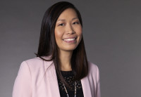 Audible's CFO Cynthia Chu stands against a grey background in a black eyelet shirt and pink blazer.