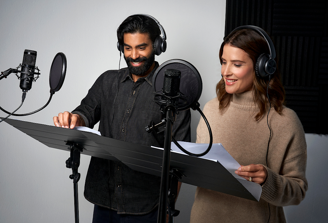 A photo of Cobie Smulders and Raymond Ablack in the studio. They're wearing headphones and reading lines from a script. 