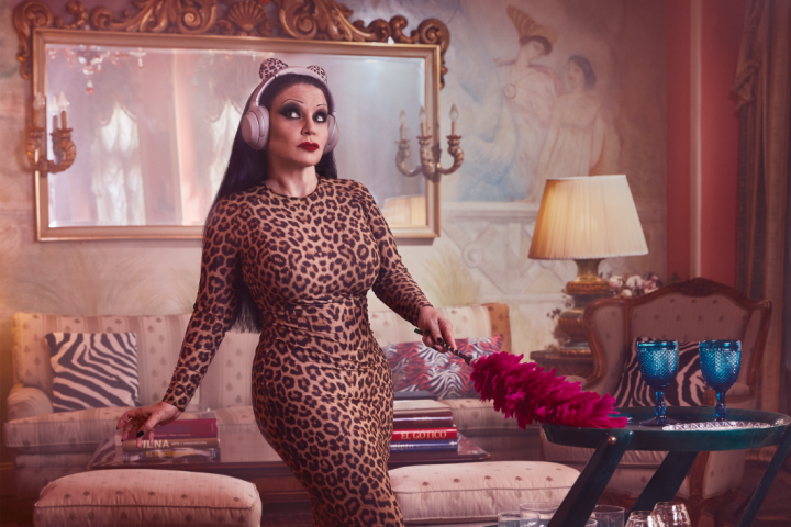 A woman stands in her opulently decorated living room wearing a leopard print dress, leopard ears on her head along with pink earphones and holds a pink duster in her hand. She is listening intently to Audible while doing housework.