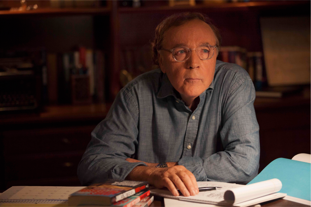 The best-selling author James Patterson sits behind his desk staring off camera. His arms are crossed and propped up on the desk. A pad of paper sits on his desk, a pen lying across it by his hand. 