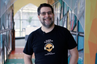 Oren Levin stands straight at the camera smiling and wearing a black shirt that features an alien wearing headphones and the words "Audible Engineering" above it. Behind him is a colorful backdrop of Audible's lobby at its Newark headquarters.