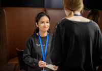 During a Women@Audible event, two employees talk to each other. The back of one of the women is facing the camera and we see beyond her to the woman facing her, who is listening and starting to smile. They both wear dark crew neck shirts and blue lanyards around their necks with their employee IDs.
