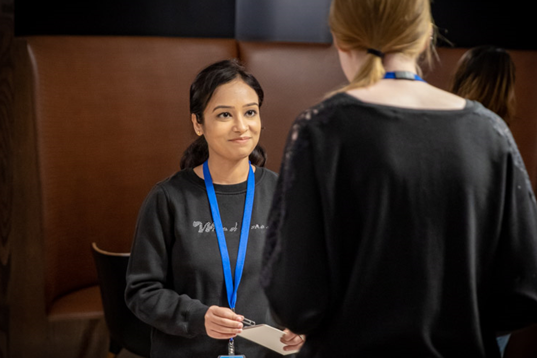 During a Women@Audible event, two employees talk to each other. The back of one of the women is facing the camera and we see beyond her to the woman facing her, who is listening and starting to smile. They both wear dark crew neck shirts and blue lanyards around their necks with their employee IDs.