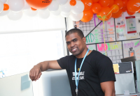 Sanchez Brown smiles directly at the camera with one arm leaning up on his desk at the Audible headquarters in Newark. Orange and white balloons with Audible logos displayed surround him.