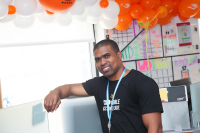 Sanchez Brown smiles directly at the camera with one arm leaning up on his desk at the Audible headquarters in Newark. Orange and white balloons with Audible logos displayed surround him.