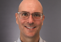 A close cropped headshot of Audible CTO Tim Martin with his face, smiling, shown against a grey background. He is wearing grey framed glasses and a grey and white checked button down shirt.