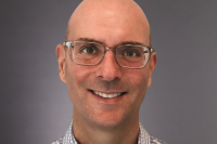 A close cropped headshot of Audible CTO Tim Martin with his face, smiling, shown against a grey background. He is wearing grey framed glasses and a grey and white checked button down shirt.