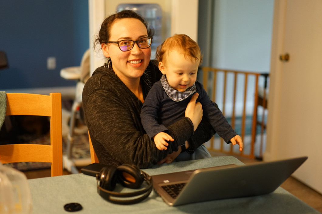 A woman sits at her kitchen table, her son, a toddler, held in her lap, a laptop open on the table in front of her.