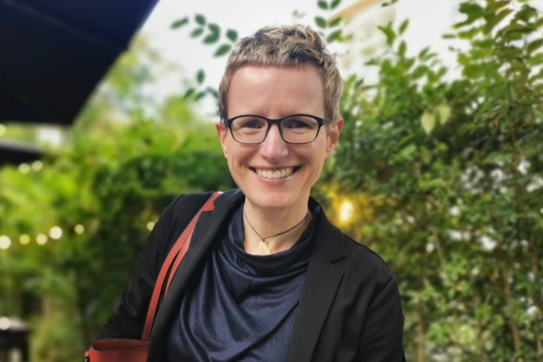 Audible employee Sibylle Schwarz stands in the outdoor patio of a restaurant with greenery behind her, black table umbrellas overhead, and lights strung across the background. She is looking directly to the camera and smiling. She has short cropped blond hair and is wearing dark framed glasses, a navy shirt and a black blazer. She has a red purse slung over her shoulder.