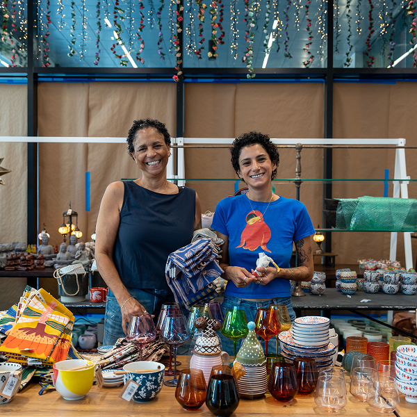 Two people stand behind a table piled with goods for sale.