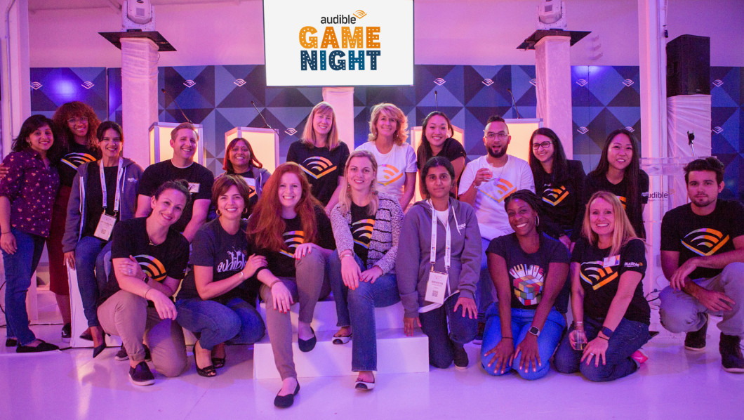 A large group of women wearing t-shirts with Audible's logo pose together before an Audible Game Night sign.
