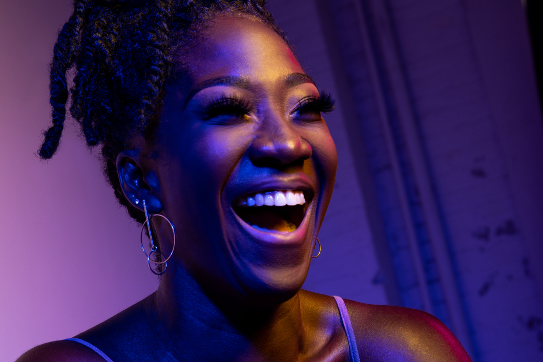 Amber Iman smiles and is glowing with a purple hue coming from a spotlight.