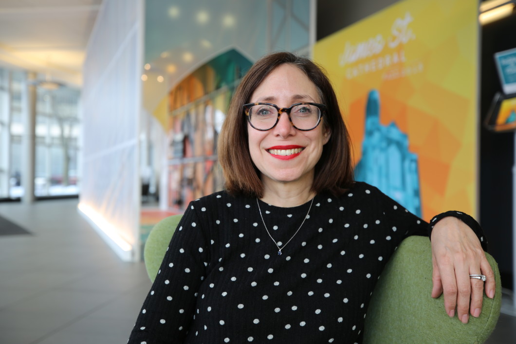 Kristin Lang, Audible’s Senior Director of Acquisitions and Content Partnerships, sits on a green chair in the lobby of Audible's headquarters. She is staring straight at the camera and wearing glasses and a black shirt with white polka dots. 