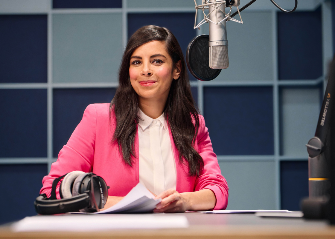 Collien Ulmen-Fernandes sits in a recording studio in front of a microphone, smiling to camera.