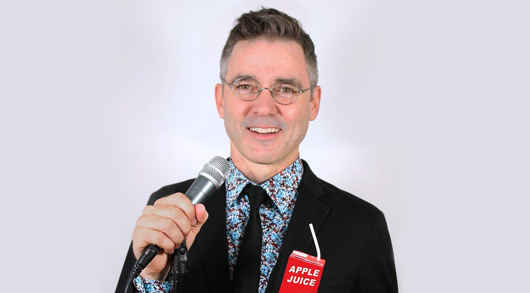Comedian Billy Kelly, wearing wire-framed glasses, a blue and purple patterned shirt, black tie and suit jacket, stands holding a microphone in his right hand and a apple juice box with straw in his left hand.