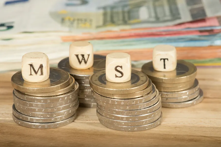 Hier sehen Sie ein Foto von Münzgeld, welches aufeinander gestapelt ist. Im Hintergrund liegen Scheine. Auf den Münz-Türmen liegen Holzperlen auf denen die Buchstaben "M", "W", "S" und "T" zu sehen sind.