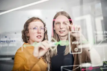 Hier sehen Sie zwei Mitarbeiterinnen von Homeday vor einer Glasscheibe.