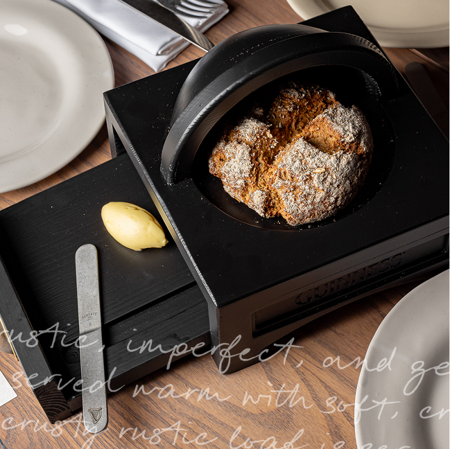 A round loaf of bread in a black lidded dish, with a pat of butter and a knife on a black tray, set on a wooden table with white plates and cutlery. 
