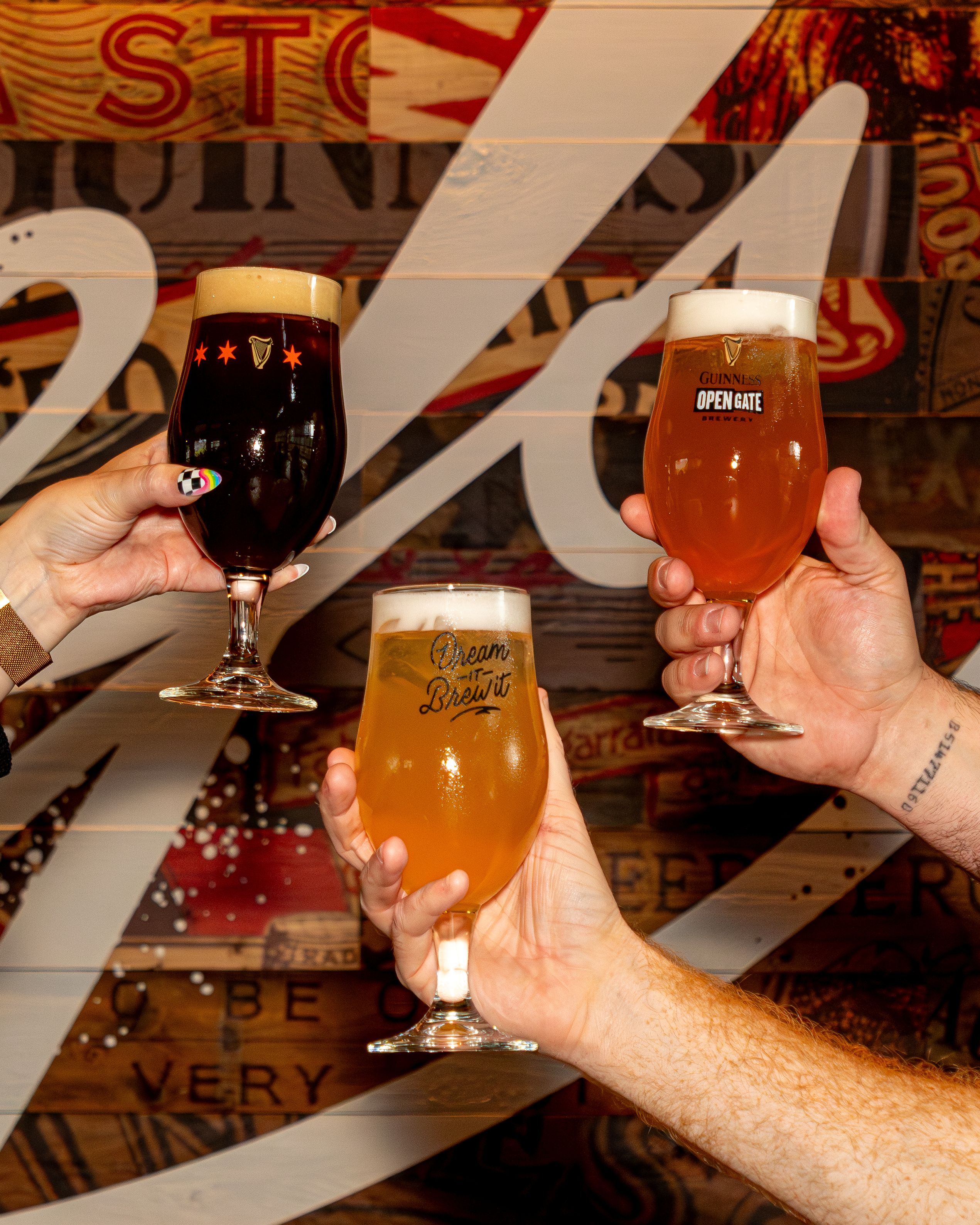 Three hands hold different Guinness beers in stemmed glasses against a rustic wooden background with painted letters and graphics.