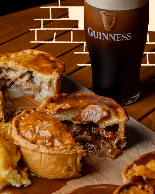 Golden-brown pies, some open with savory fillings, on a wooden surface next to a pint of Guinness. White brick pattern in the background.
